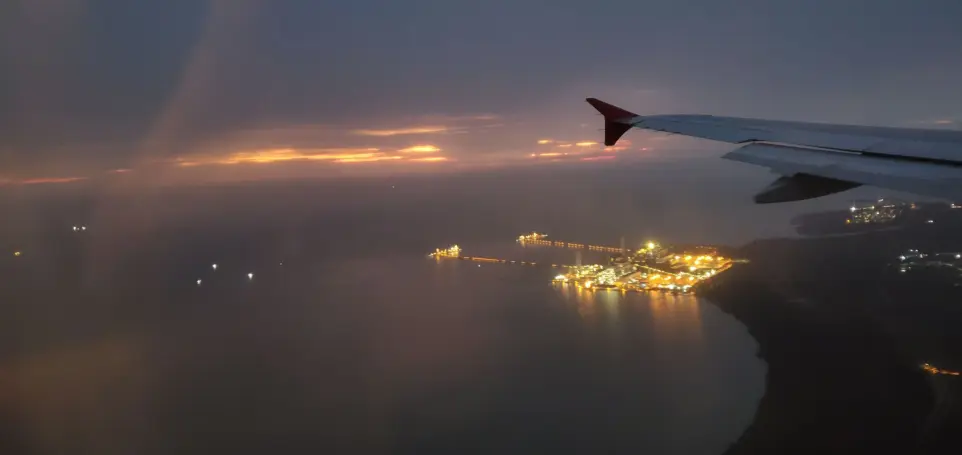 from airplane window with wing in foreground, orange sunset horizon, and illuminated coastline over calm ocean